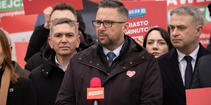 Polityk w brązowym płaszczu i okularach przemawia do mikrofonu Lewica podczas wiecu wyborczego, otoczony innymi kandydatami i czerwonymi plakatami w tle.