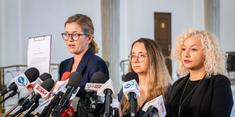 Trzy kobiety podczas konferencji prasowej z licznymi mikrofonami; jedna trzyma kartkę z dokumentem, omawiając projekt Lewicy dot. bezreceptowej tabletki „dzień po”.