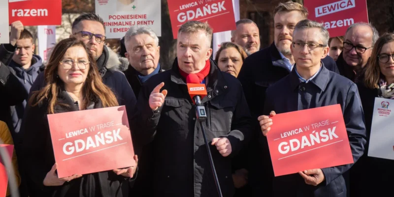 Grupa zwolenników Lewicy podczas wiecu, mężczyzna przemawia do mikrofonu na tle czerwonych plakatów z napisami LEWICA i GDANSK.