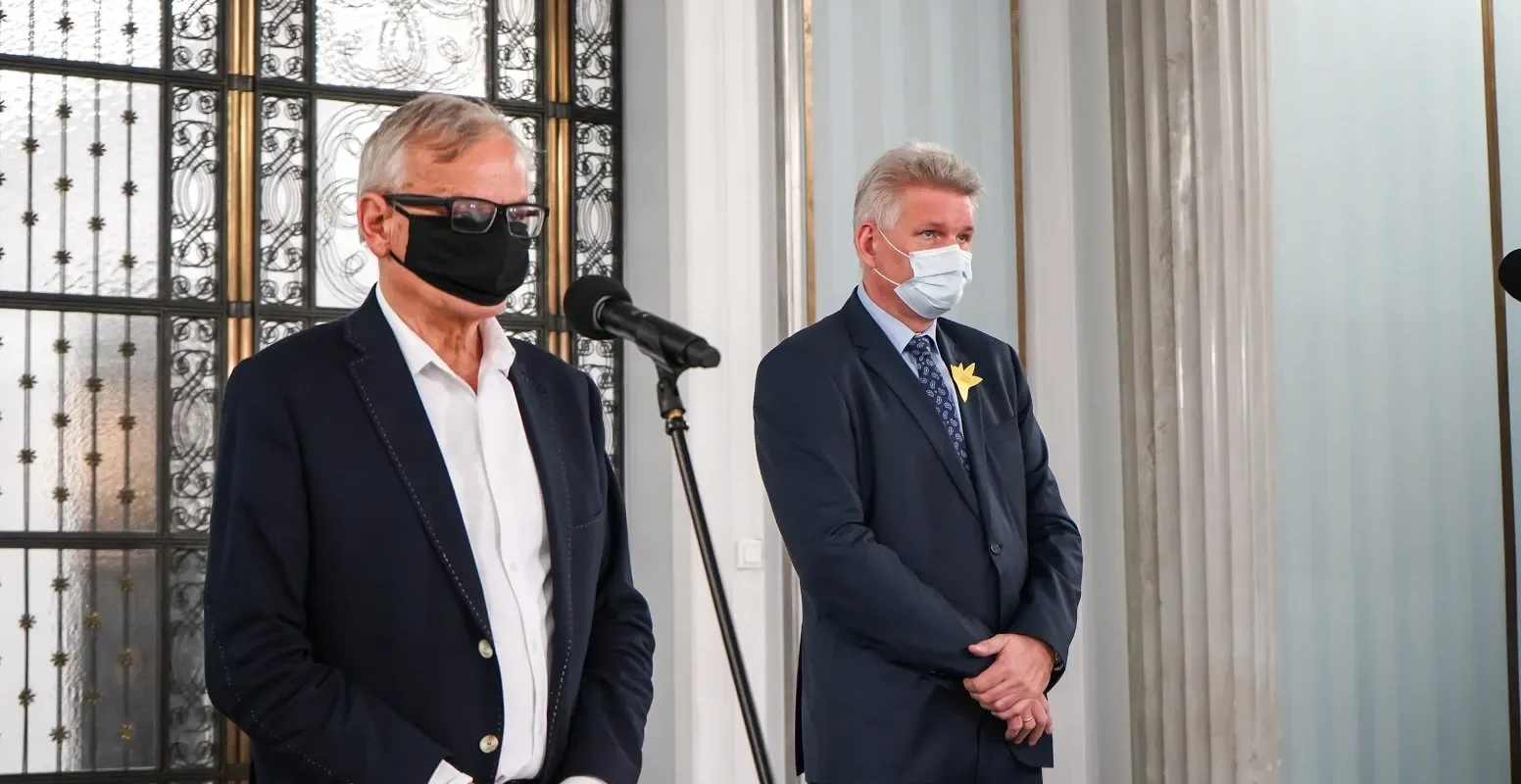 Dwóch mężczyzn w garniturach stoi przy mikrofonach podczas konferencji, jeden ma czarną maseczkę i okulary, drugi białą maseczkę i żółty kwiat na klapie, a w tle widać ozdobne drzwi i kolumny.