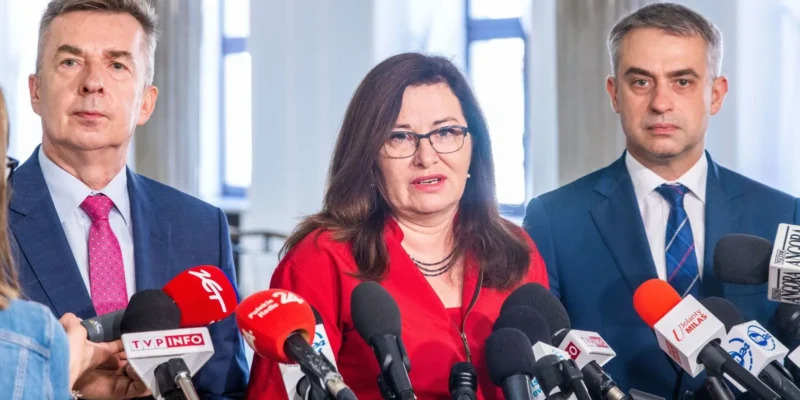 Trzy osoby w garniturach stoją przy licznych mikrofonach na konferencji prasowej; pośrodku kobieta w czerwonej bluzce przemawia do reporterów.