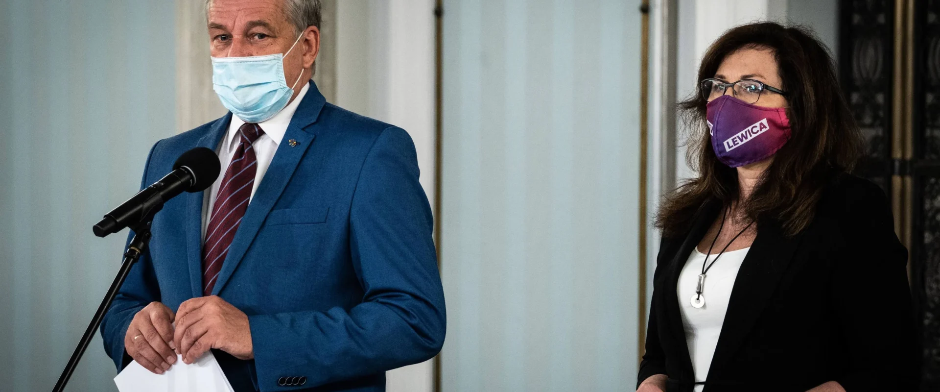 Dwóch polityków podczas konferencji prasowej stojących przy mikrofonach; po lewej mężczyzna w niebieskim garniturze i maseczce, po prawej kobieta w czarnym żakiecie i fioletowej masce z napisem Lewica.