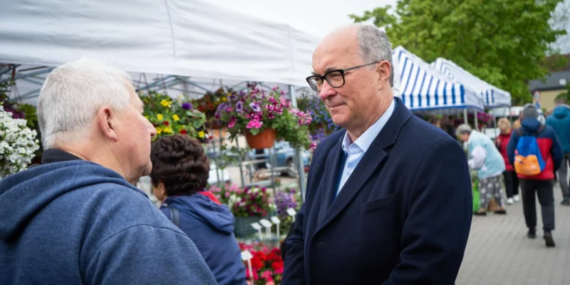 Dwóch mężczyzn rozmawia na targowisku z kwiatami, stojąc obok stoisk pod białymi i paskowanymi namiotami.