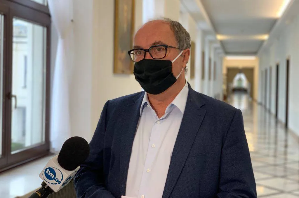 Mężczyzna w garniturze i białej koszuli, noszący czarną maseczkę i okulary, udziela wywiadu reporterowi z mikrofonem TVN24 w długim, jasnym korytarzu.