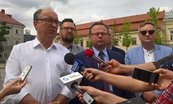 Kilku mężczyzn w garniturach stoi na zewnątrz podczas konferencji prasowej, otoczonych mikrofonami i dyktafonami dziennikarzy.