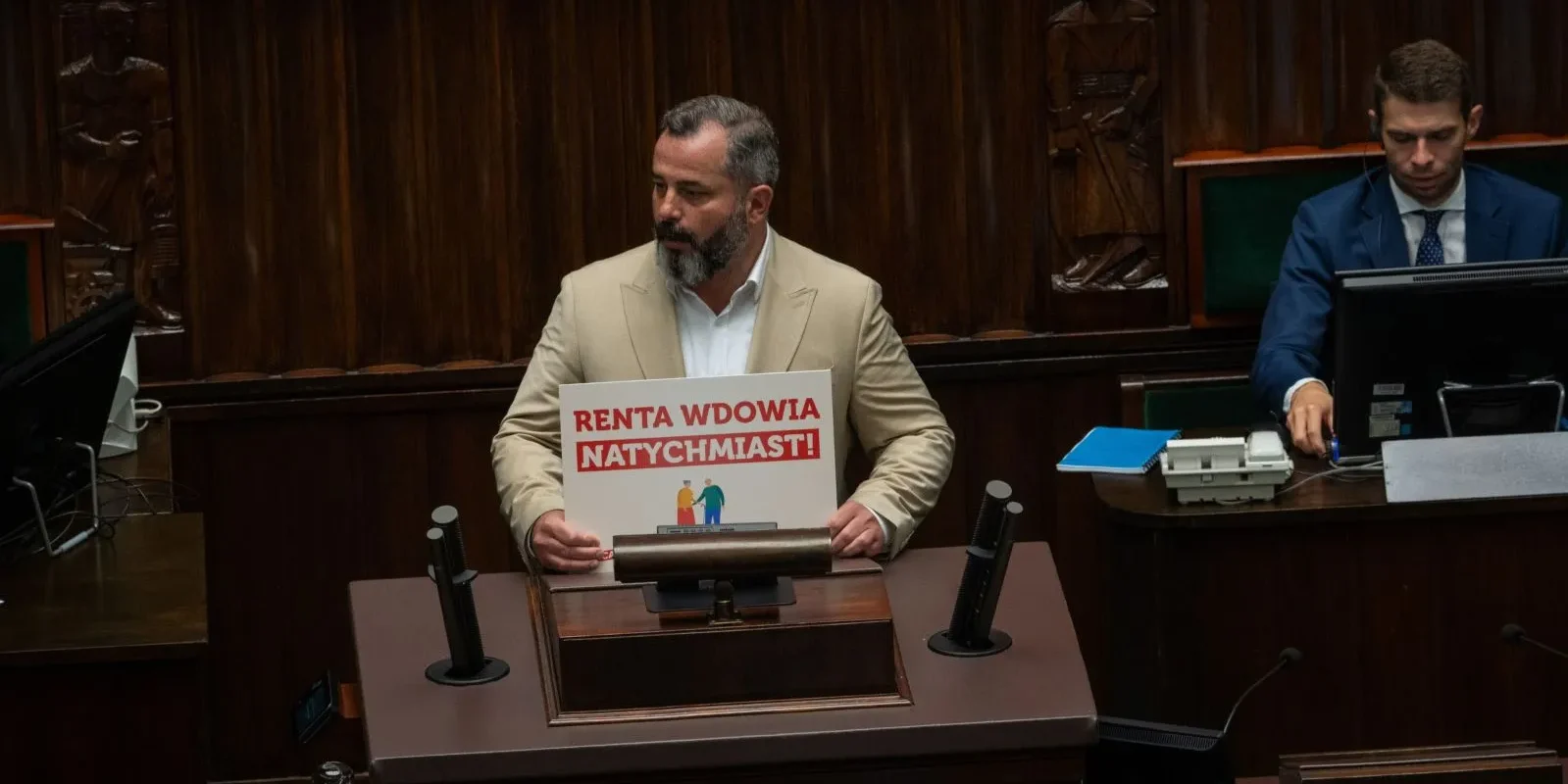 Mężczyzna w jasnym garniturze stoi przy mównicy Sejmu i trzyma plakat z napisem „Renta wdowia natychmiast!”.