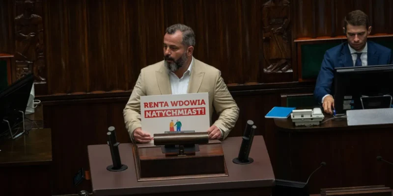Mężczyzna w jasnym garniturze stoi przy mównicy Sejmu i trzyma plakat z napisem „Renta wdowia natychmiast!”.
