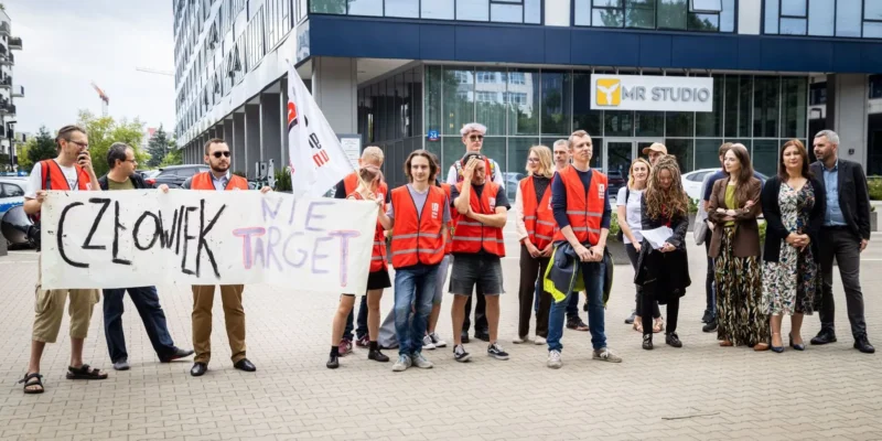 Grupa demonstrantów w pomarańczowych kamizelkach stoi przed biurowcem MR Studio, trzymając baner domagający się zakazu zwalniania związkowców do czasu ogłoszenia wyroku sądu.