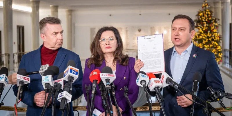 Trójka polityków stoi podczas konferencji prasowej przed mikrofonami, kobieta w fioletowym żakiecie trzyma dokument, po obu stronach stoją mężczyźni, a w tle widoczna choinka.