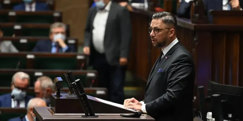 Mężczyzna w garniturze przemawia z mównicy podczas posiedzenia w sali plenarniej, trzymając dokumenty, podczas gdy w tle siedzący uczestnicy w maseczkach obserwują wydarzenie.