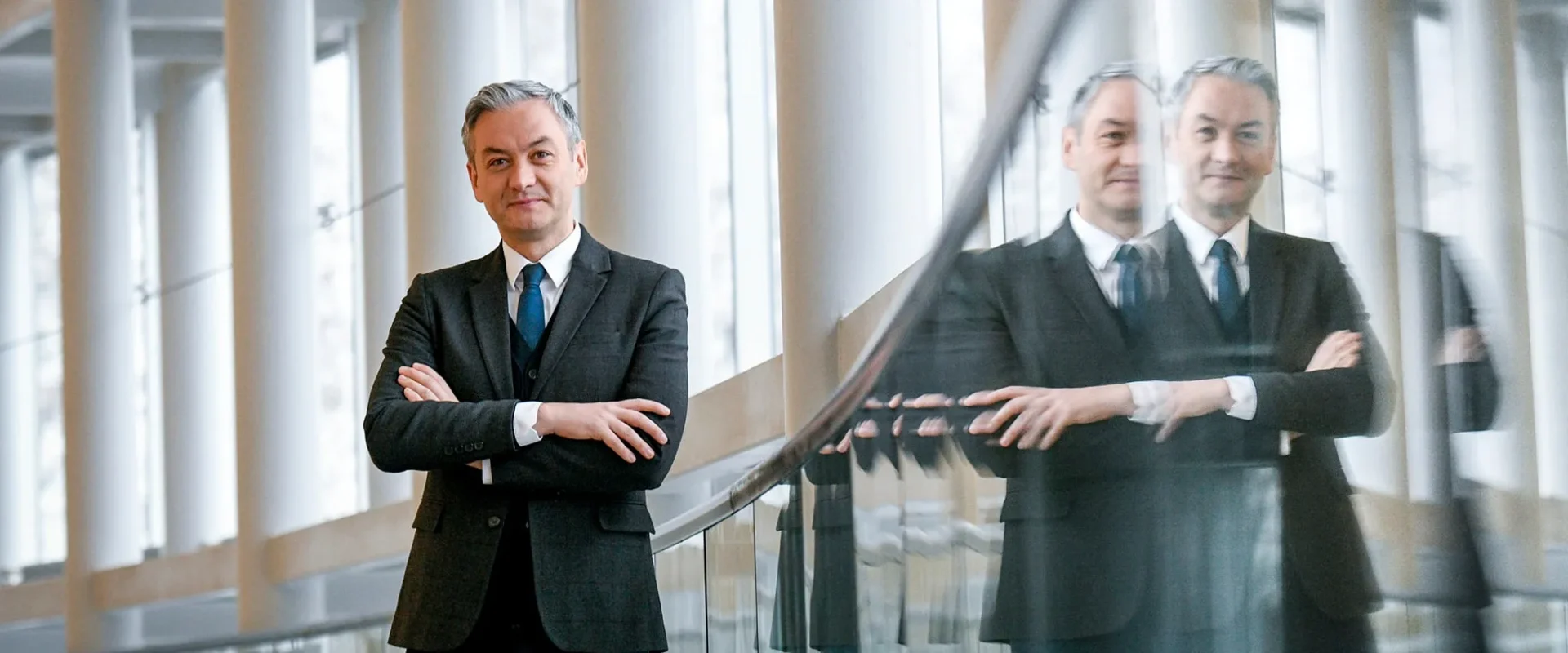 Mężczyzna w garniturze stoi w eleganckim wnętrzu, z założonymi rękami, a w tle widać jego odbicie w szklanej balustradzie.