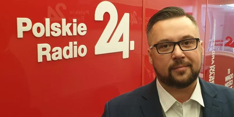 Mężczyzna w garniturze i okularach stoi przed czerwonym tłem z logo Polskie Radio 24.