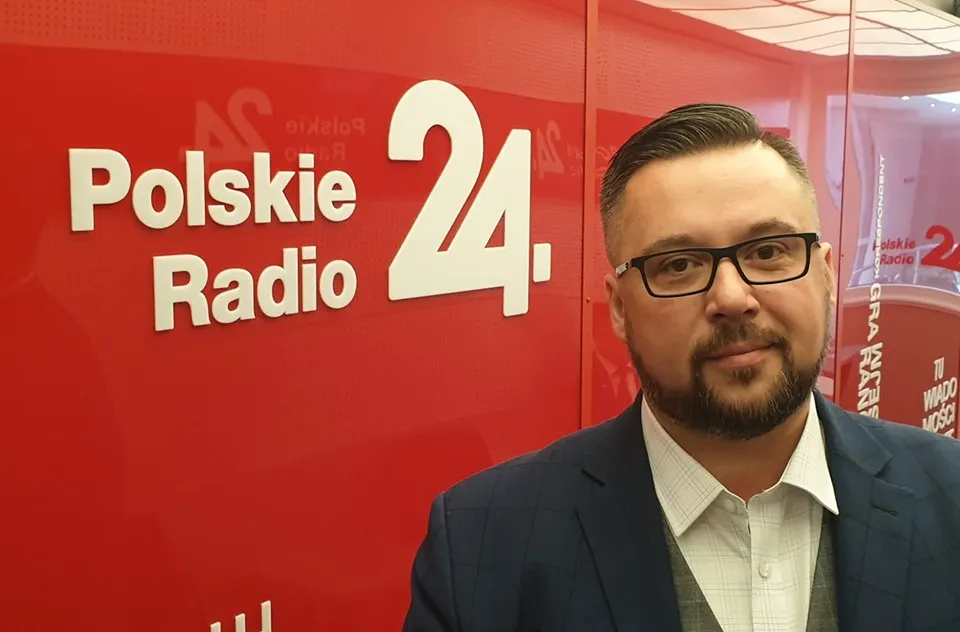 Mężczyzna w garniturze i okularach stoi przed czerwonym tłem z logo Polskie Radio 24.