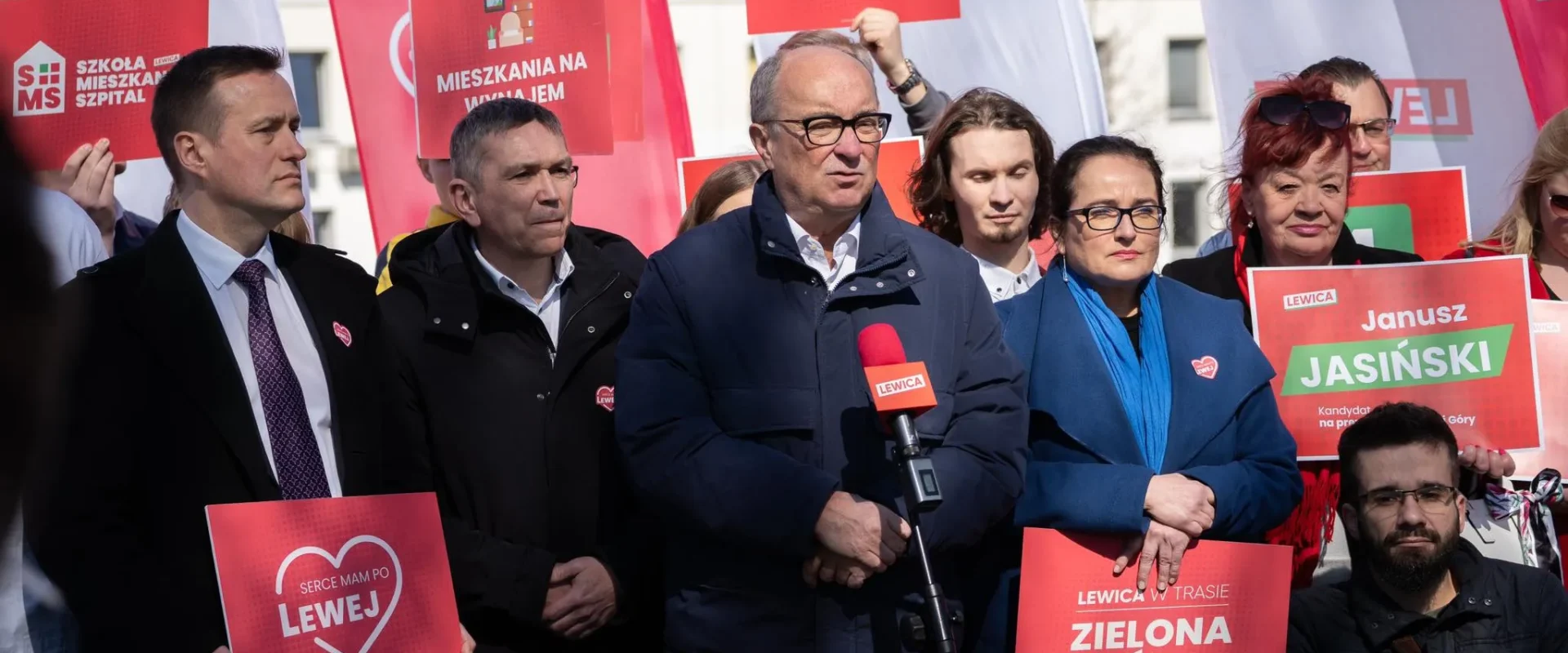 Grupa polityków Lewicy na wiecu, stojąca przed czerwonymi plakatami i banerami, jeden z nich przemawia do mikrofonu.