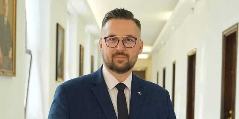 Mężczyzna w granatowym garniturze i okularach stoi w korytarzu, uśmiechnięty, z przypinką w barwach flagi na klapie.