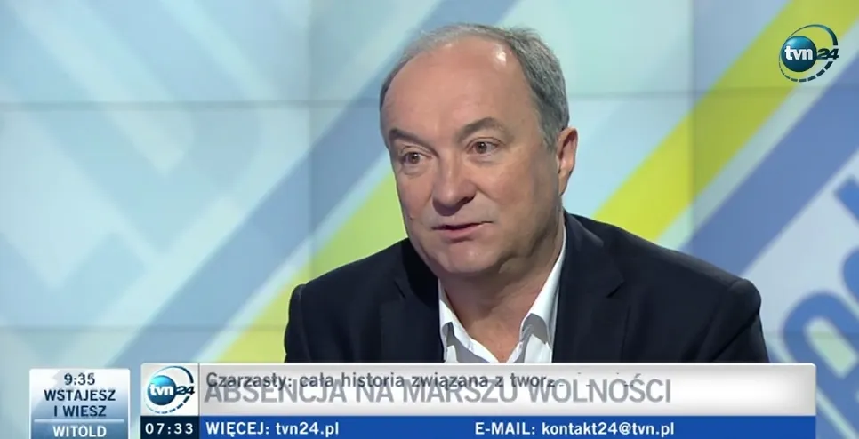 Mężczyzna w marynarce i białej koszuli rozmawia w studiu telewizyjnym, z widocznym logo TVN24 i paskiem informacyjnym u dołu ekranu.
