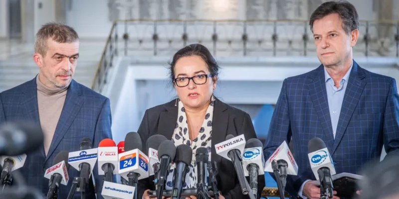 Trzy osoby w garniturach stojące za licznymi mikrofonami podczas konferencji prasowej.