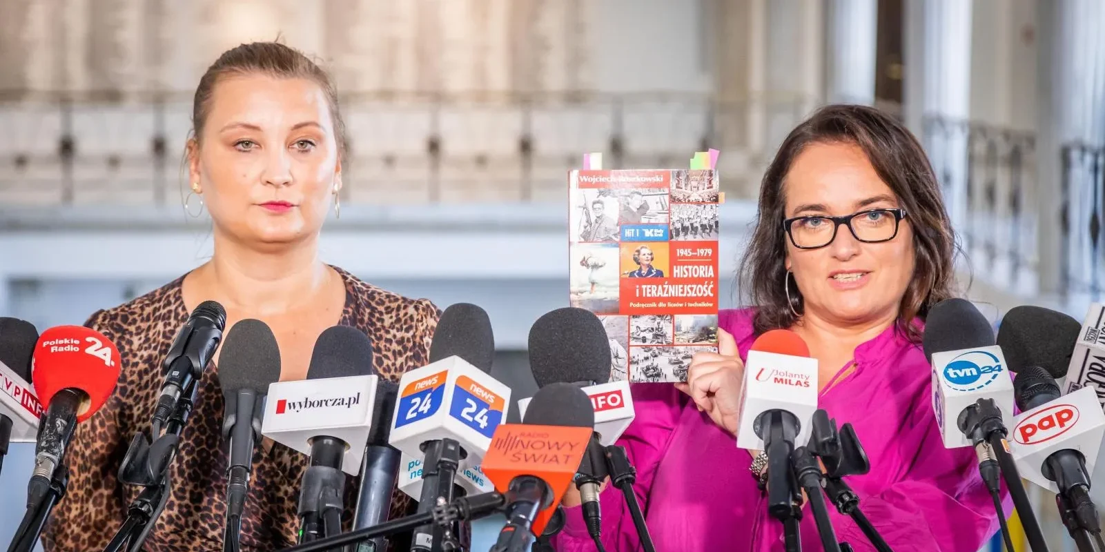 Na zdjęciu przedstawiono dwie kobiety podczas konferencji prasowej, jedna z nich trzyma książkę zatytułowaną "Historia i teraźniejszość", a wokół nich znajduje się wiele mikrofonów mediów.