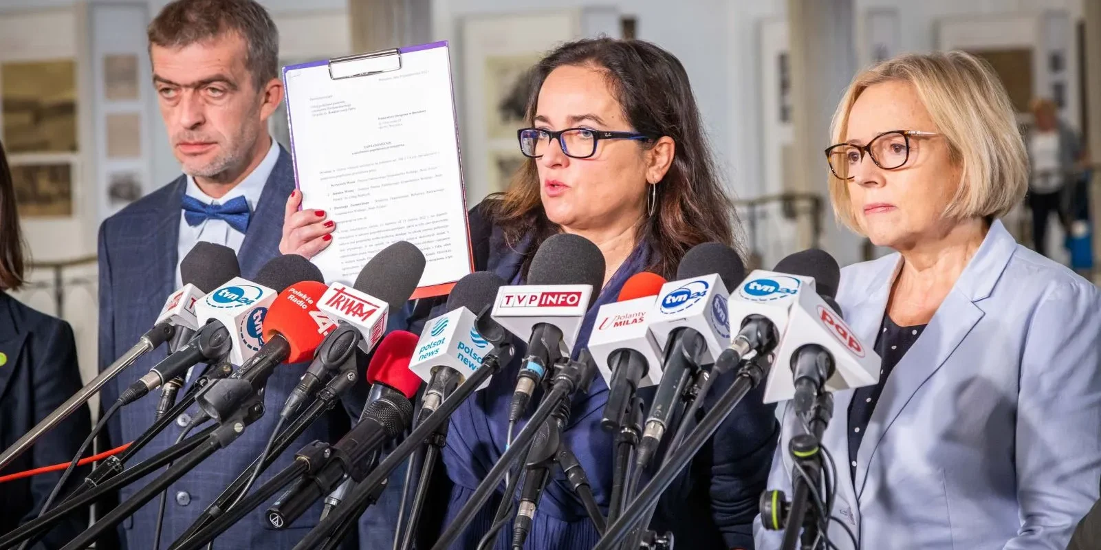 Trzech mówiących ludzi stoi przed mikrofonami i prezentuje dokument na konferencji prasowej.