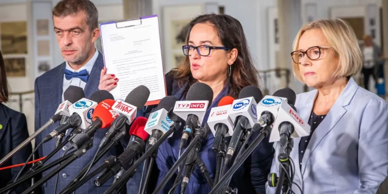 Trzech mówiących ludzi stoi przed mikrofonami i prezentuje dokument na konferencji prasowej.