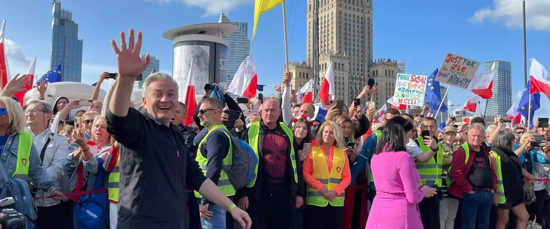 Tłum uczestników marszu na ulicy z flagami i żółtymi kamizelkami, na pierwszym planie mężczyzna w czarnej kurtce macha ręką, a w tle widać wysokie budynki i błękitne niebo.