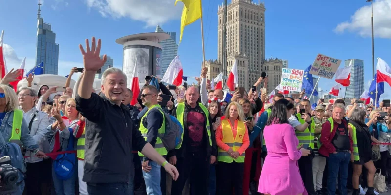 Tłum uczestników marszu na ulicy z flagami i żółtymi kamizelkami, na pierwszym planie mężczyzna w czarnej kurtce macha ręką, a w tle widać wysokie budynki i błękitne niebo.