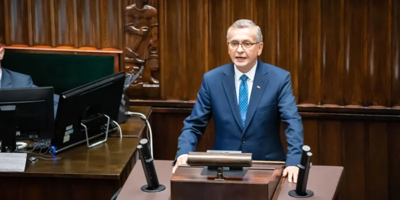 Mężczyzna w granatowym garniturze i niebieskim krawacie stoi za mównicą w sejmowej sali obrad i wygłasza przemówienie.