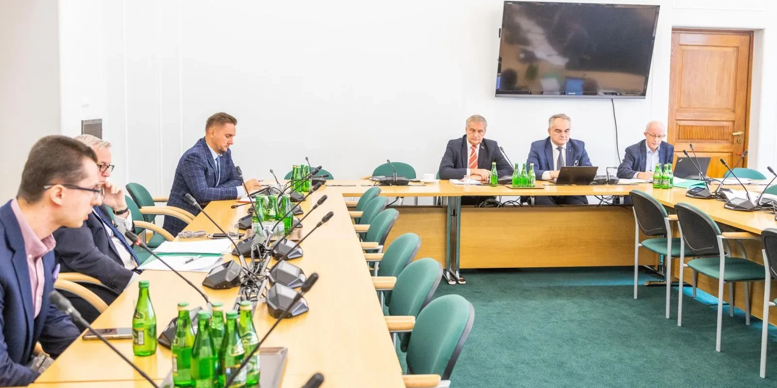 Na zdjęciu widać grupę ludzi siedzących przy stole w sali konferencyjnej, z butelkami wody i sprzętem do nagrań.