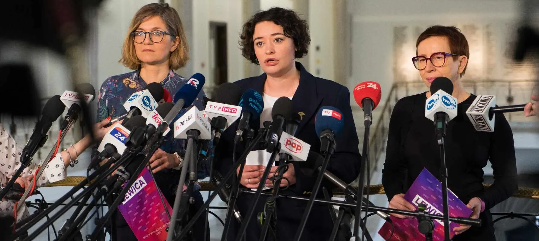 Trzy kobiety z Lewicy stoją na konferencji prasowej, otoczone licznymi mikrofonami, i prezentują projekt małej ustawy schronowej.