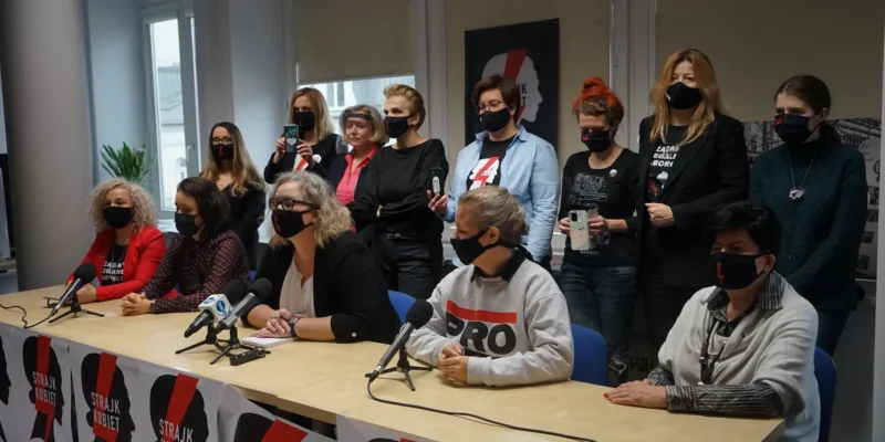 Konferencja prasowa Komitetu Inicjatywy Ustawodawczej dotycząca liberalizacji prawa aborcyjnego, edukacji seksualnej i dostępu do antykoncepcji; przy stole z mikrofonami siedzą prowadzący, a za nimi stoi grupa aktywistek i aktywistów w maseczkach, z banerami Strajk Kobiet w tle.