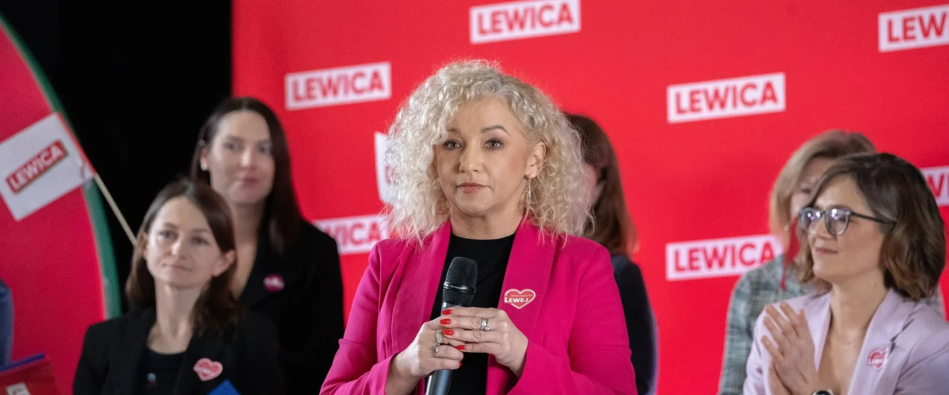 Kobieta o blond lokach w różowej marynarce trzyma mikrofon i przemawia podczas wydarzenia Lewicy, na tle czerwonych banerów z napisem LEWICA, podczas gdy inni uczestnicy ją słuchają i klaskają.
