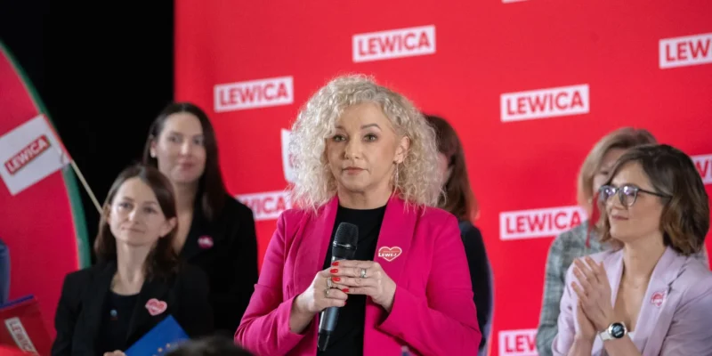 Kobieta o blond lokach w różowej marynarce trzyma mikrofon i przemawia podczas wydarzenia Lewicy, na tle czerwonych banerów z napisem LEWICA, podczas gdy inni uczestnicy ją słuchają i klaskają.