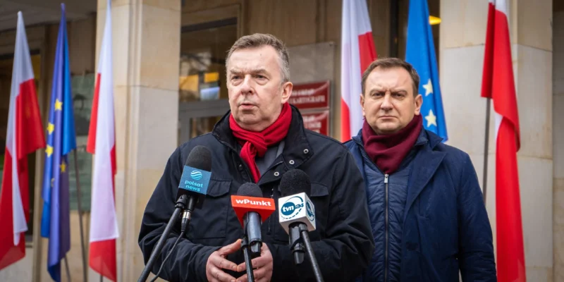 Na zdjęciu widać dwóch mężczyzn przemawiających podczas konferencji prasowej, stojących przed grupą flag, w tym flagami Polski i Unii Europejskiej.