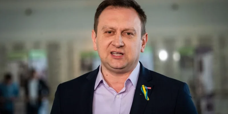 Mężczyzna w garniturze z odsłoniętymi ramionami mówi do kamery, mając na sobie przypinkę w kolorach flagi Ukrainy.