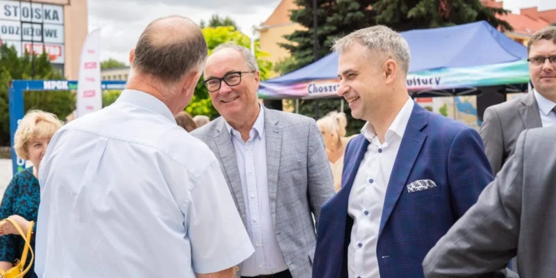 Trzech mężczyzn w garniturach rozmawia i uśmiecha się podczas plenerowego wydarzenia, w tle namioty i inne osoby.