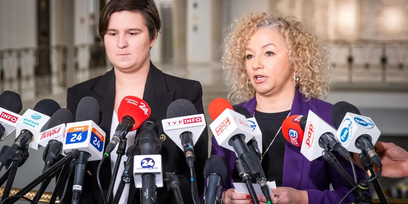 Dwie kobiety mówiące do mediów podczas konferencji prasowej, otoczone licznymi mikrofonami ustawionymi na statywach w holu.