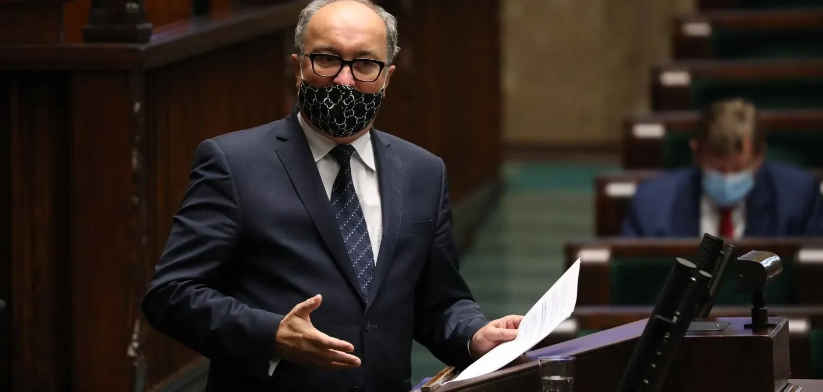 Polityk w ciemnym garniturze i maseczce z wzorem stoi przy mównicy w sali plenarnej, trzyma kartkę i gestykuluje.