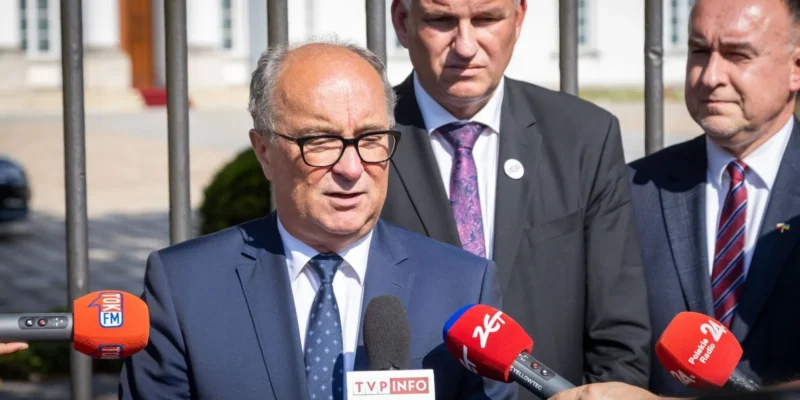 Mężczyzna w granatowym garniturze i okularach udziela wywiadu przed mikrofonami dziennikarzy podczas konferencji prasowej na zewnątrz.