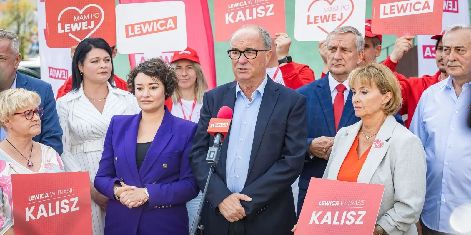 Grupa działaczy Lewicy podczas konferencji prasowej trzyma plakaty z napisem „LEWICA” i „KALISZ”, stojąc wokół mówcy przy mikrofonie.
