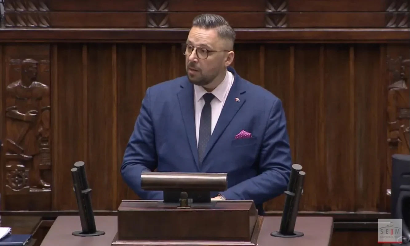 Mężczyzna w garniturze przemawia z mównicy w sejmie, z tłem ciemnego drewna i rzeźbami.