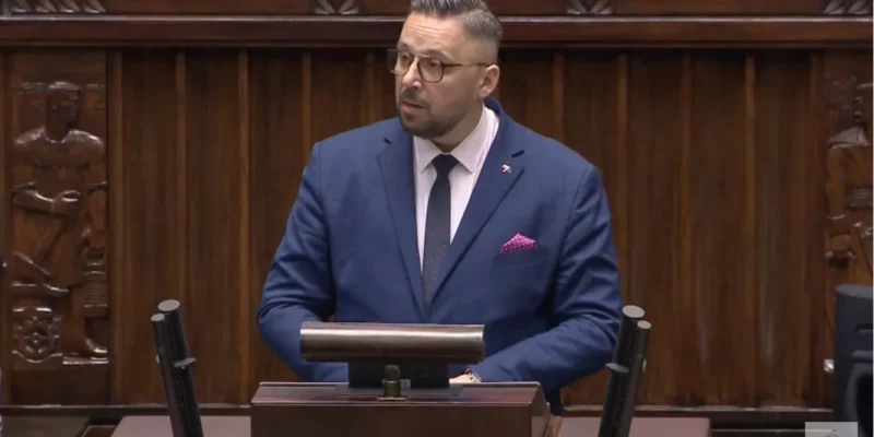 Mężczyzna w garniturze przemawia z mównicy w sejmie, z tłem ciemnego drewna i rzeźbami.