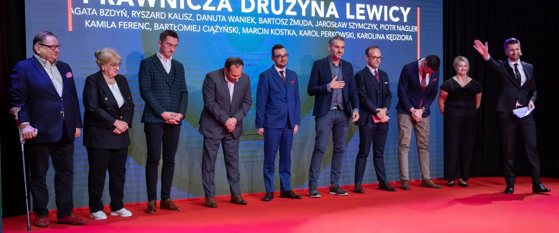 Na zdjęciu widoczna jest grupa osób na czerwonym dywanie, przedstawiająca prawników związanych z drużyną Lewicy, stojących w linii, z jedną osobą na podium.