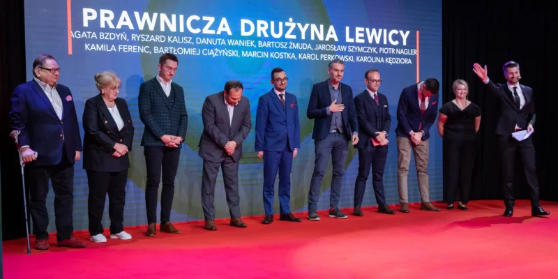 Na zdjęciu widoczna jest grupa osób na czerwonym dywanie, przedstawiająca prawników związanych z drużyną Lewicy, stojących w linii, z jedną osobą na podium.