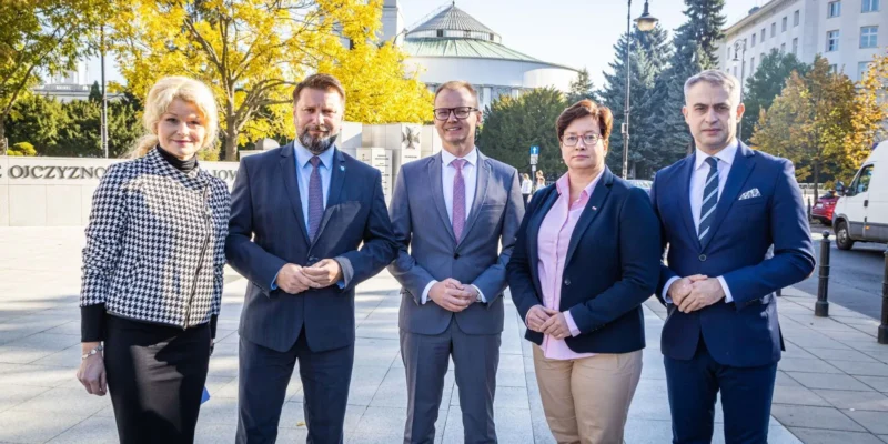 Na zdjęciu widoczna jest grupa pięciu osób w formalnych strojach, stojących na tle budynku, w słoneczny dzień, w otoczeniu drzew z żółtymi liśćmi.