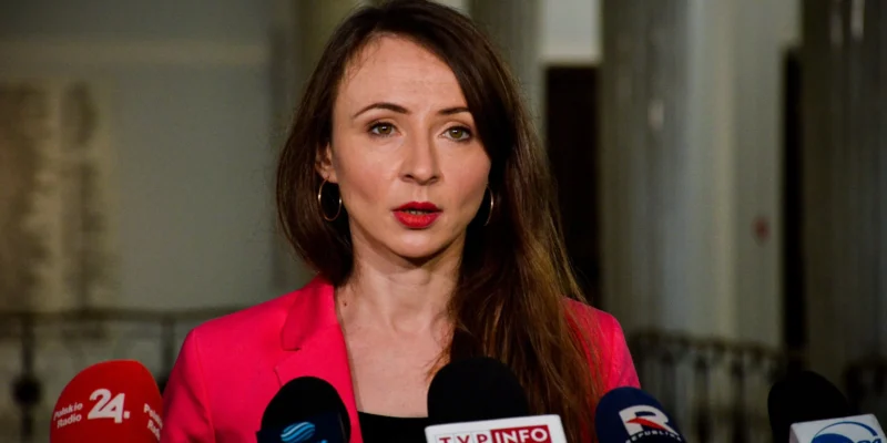 Kobieta w różowej marynarce przemawia do mediów podczas konferencji prasowej, stojąc przed kilkoma mikrofonami.