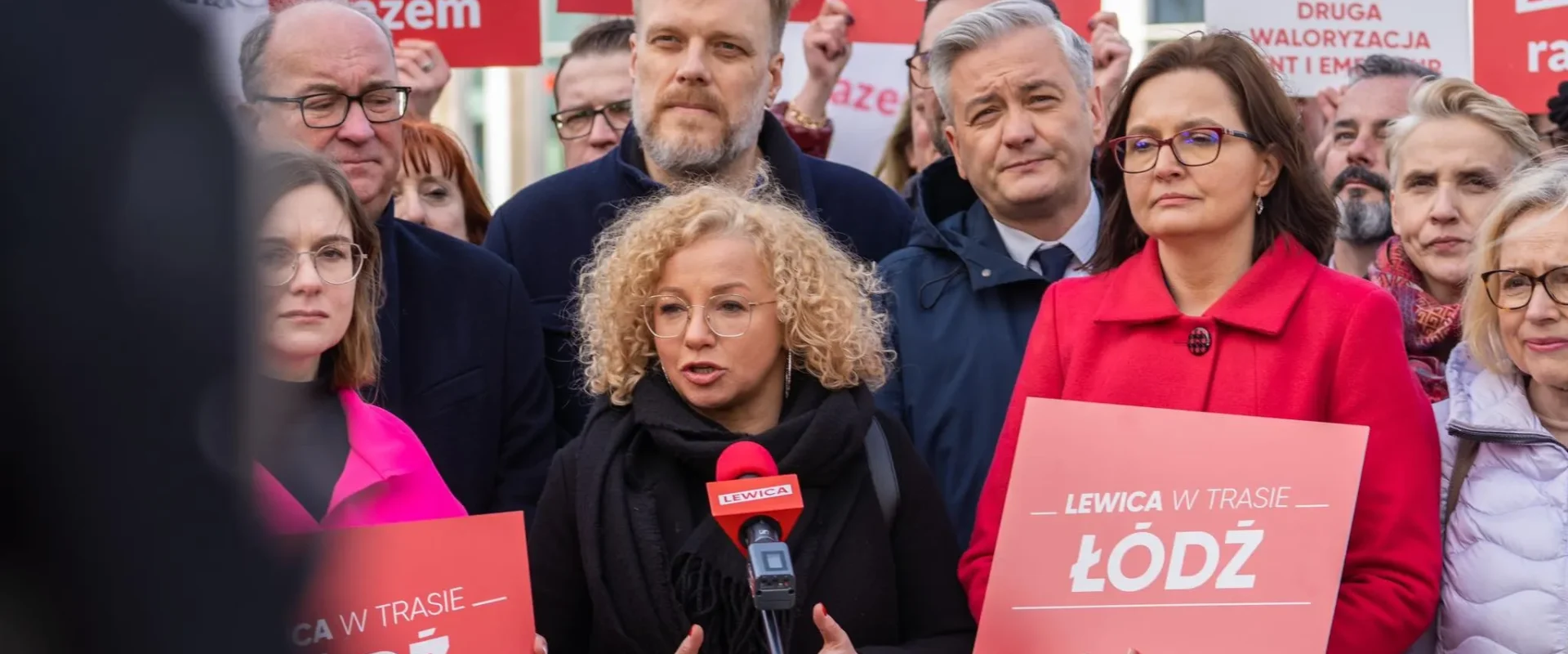 Grupa zwolenników Lewicy na wiecu w Łodzi trzyma plakaty z napisami LEWICA i ŁÓDŹ; centralnie przemawia kobieta z okrągłymi okularami do mikrofonu.