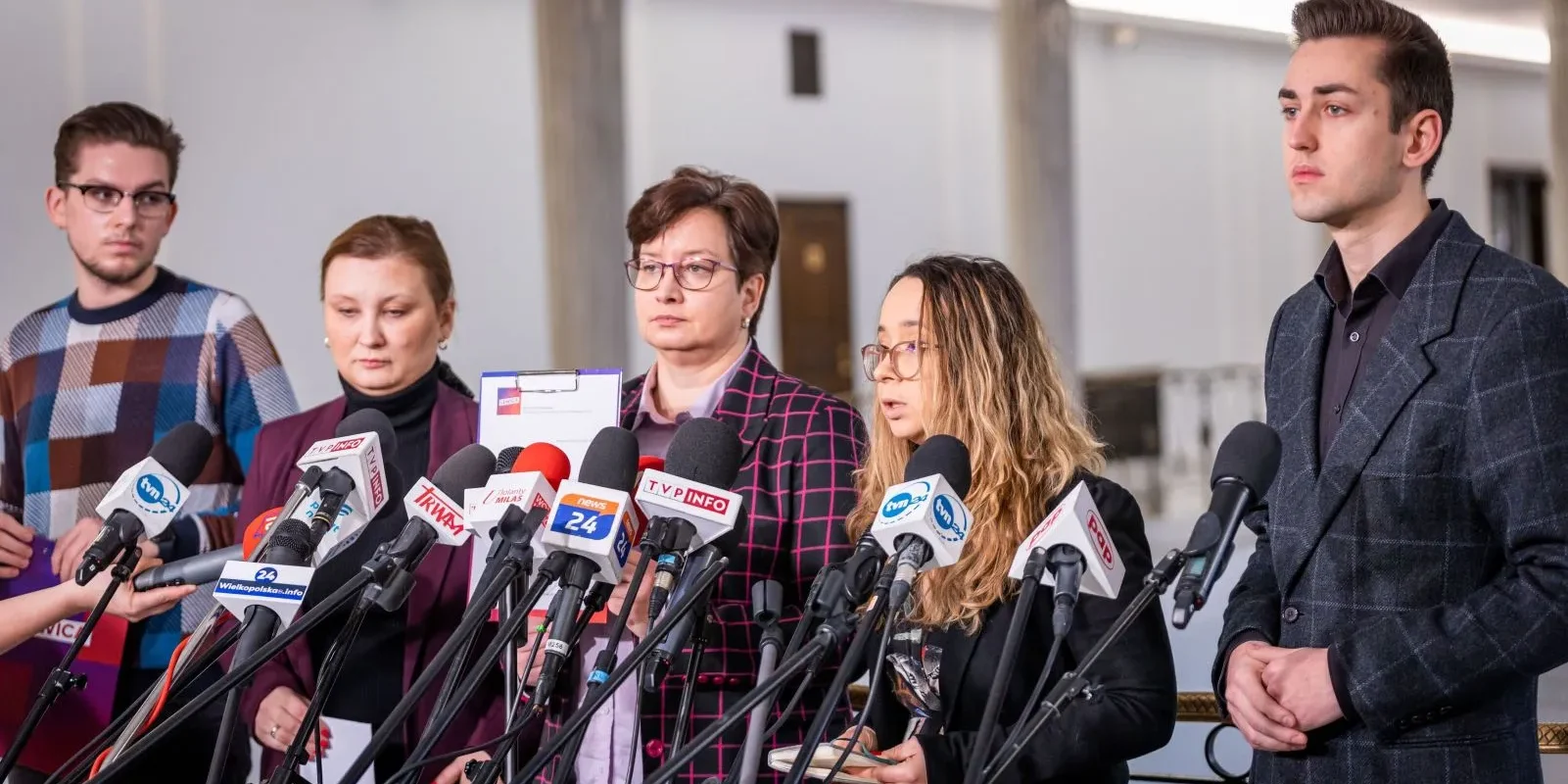 Grupa polityków i aktywistów podczas konferencji prasowej w otoczeniu licznych mikrofonów mediów.