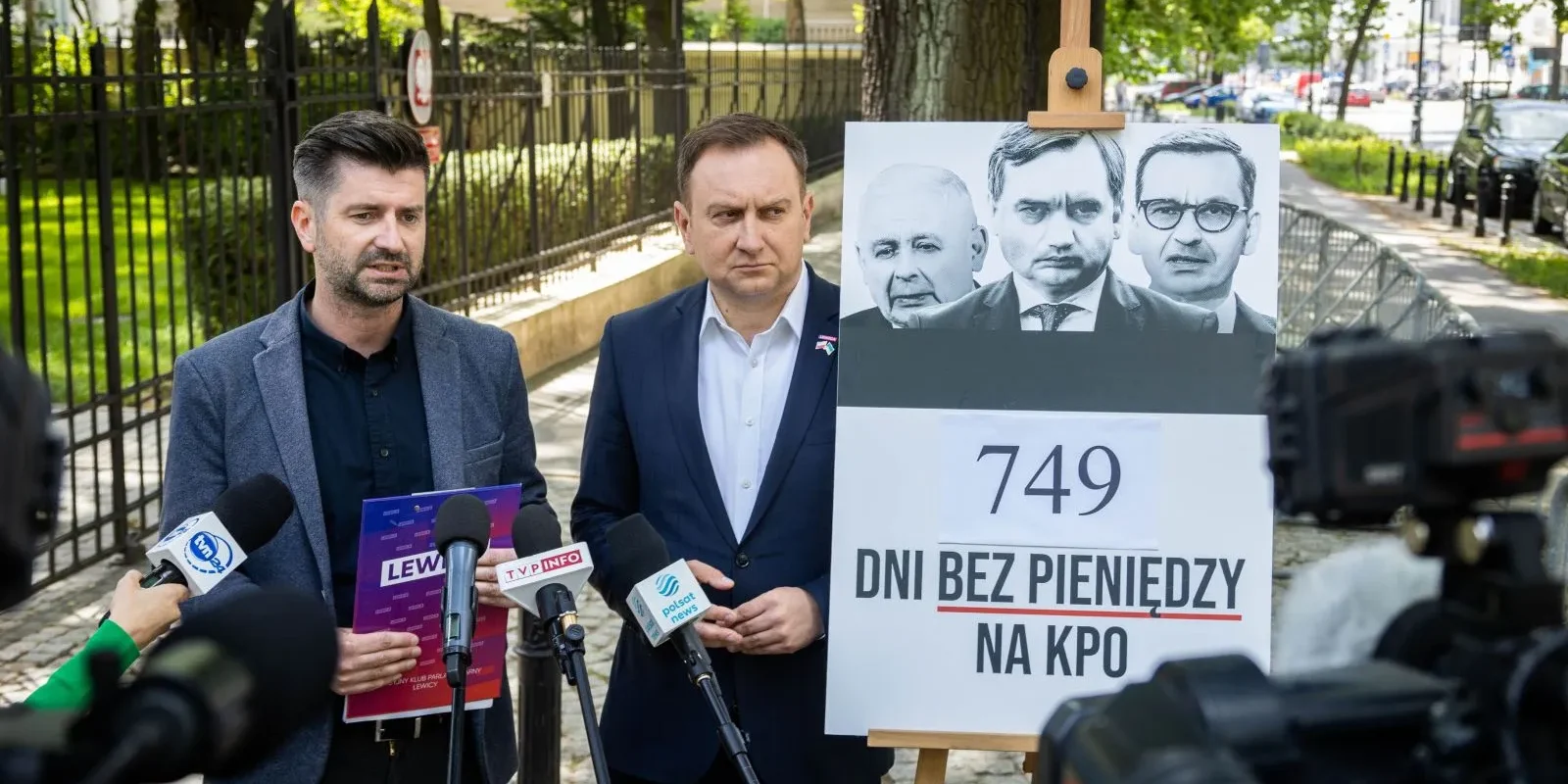 Na zdjęciu prezentują się dwaj mężczyźni podczas konferencji prasowej, obok nich znajduje się plakat z napisem "749 DNI BEZ PIENIĘDZY NA KPO" oraz zdjęciami trzech polityków.