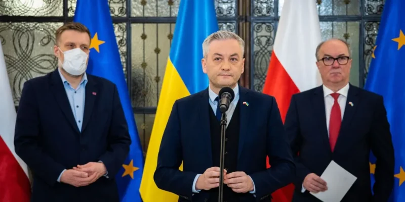 Na zdjęciu widoczna jest grupa trzech mężczyzn podczas konferencji prasowej, w tle flagi Polski, Ukrainy oraz Unii Europejskiej.