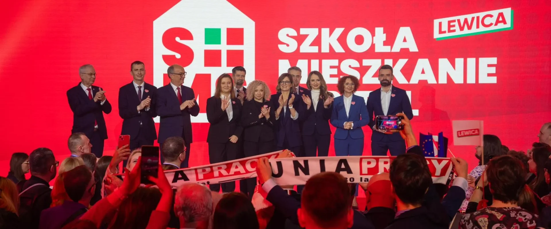 Grupa polityków Lewicy stoi na scenie podczas konwencji samorządowej, przed czerwonym tłem z napisem „SZKOŁA MIESZKANIE” i logo Lewicy, wśród wiwatującego tłumu i osób robiących zdjęcia.